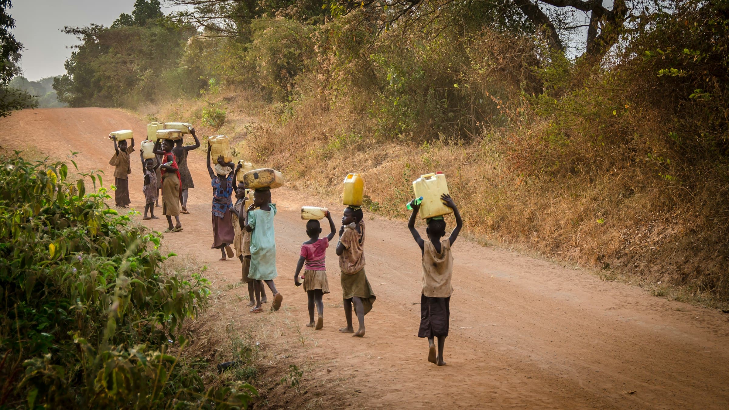 A group of people walk along a road carrying water containers in an African community.